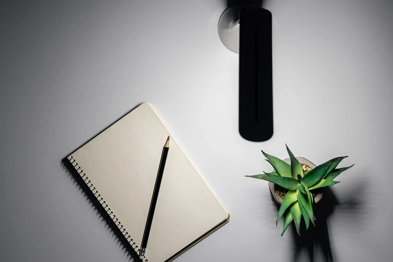 A notebook, a pencil and a small plant on a white desk, seen from above