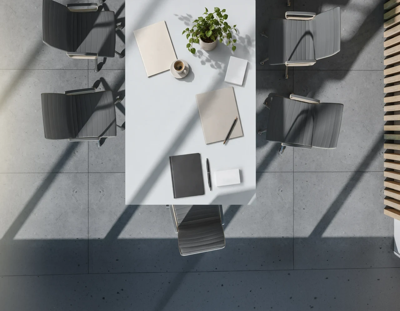 A meeting table seen from above on a concrete floor, set with notebooks, business cards, a coffee and a plant, grey chairs around it