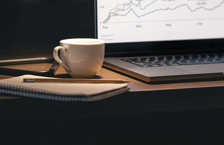A coffee cup and notebook beside a laptop showing charts