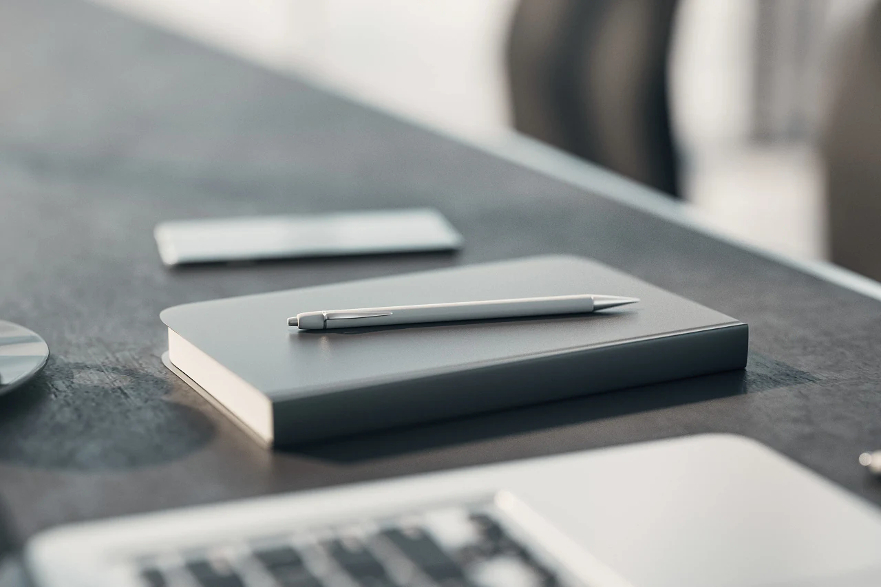 A notebook and pen on a dark desk beside a laptop